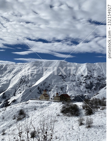 Snow-covered mountain landscape with picturesque buildings under blue sky and scattered clouds Snow-covered mountain landscape with picturesque buildings under blue sky and scattered clouds 132154927