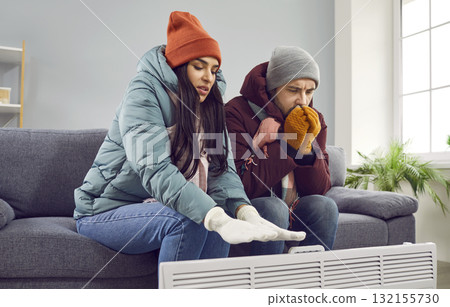 Young couple is sitting on the sofa at home in warm jacket and hat, warming their hands with heater in the living room, freezing from the cold. Heating problems, room is cold, people freezing. 132155730