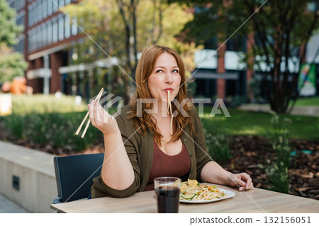 Plus size woman having lunch outdoors, eating noodles. 132156051