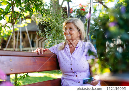 Senior woman sitting in garden, scrolling on phone. 132156095