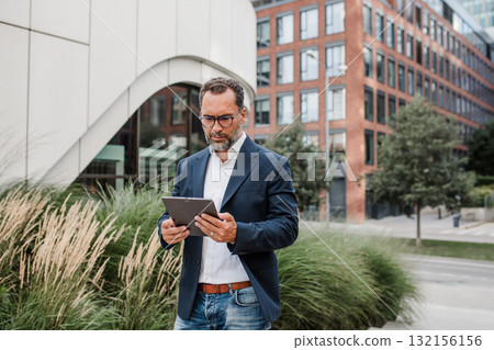 Businessman in front of office building, holding tablet and reading something. 132156156