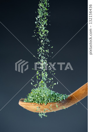 Close-up of crushed parsley or herb flakes fall from above onto a wooden spoon, ideal for culinary and food photography concepts. Close-up of crushed parsley or herb flakes fall from above onto a wooden spoon, ideal for culinary and food photography concepts. 132156436