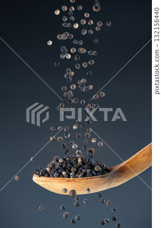 Close-up of dried black pepper corns fall from above onto a wooden spoon, perfect for gourmet food photography and spice concepts. 132156440