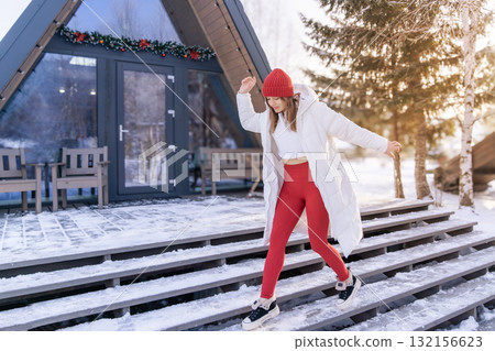 Woman walks carefully down stairs in winter attire near a holiday-themed cabin in a snowy landscape during daylight Woman walks carefully down stairs in winter attire near a holiday-themed cabin in a snowy landscape during daylight 132156623