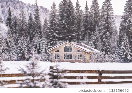 Charming cabin rests peacefully in a snowy landscape, surrounded by tall, snow-covered pine trees. Tranquil setting captures the beauty of winter in the mountains, with a wooden fence framing scene. 132156632