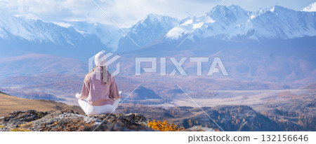 A woman sits in a meditative pose on a rock, enjoying the calming beauty of autumn mountains. The peaks are snow-capped, and the landscape is serene and tranquil. 132156646