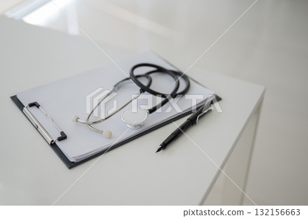 A stethoscope over clipboard with documents next to a fountain pen on white table in the white room. A stethoscope over clipboard with documents next to a fountain pen on white table in the white room. 132156663