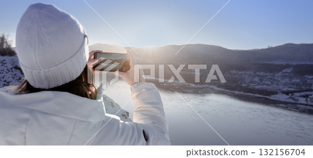 Photographing nature on a winter adventure trip - A girl in a white jacket takes pictures of a beautiful river and mountains from a high bank. rear view A person dressed in white attire is joyfully 132156704