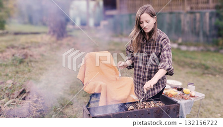 A woman in a brown shirt cooks a barbecue on the fire - she roasts mushrooms on a grill. Vegetarian picnic in autumn. 132156722
