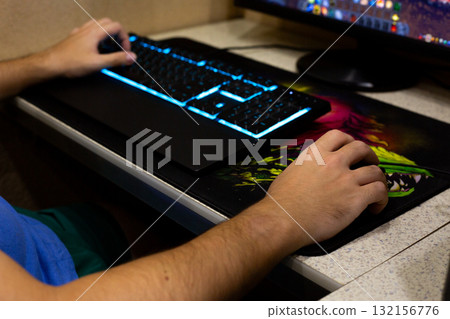 Close up of hand on gaming mouse, with backlit keyboard glowing in blue, showcasing colorful gaming mousepad. The screen displays game, highlighting the immersive experience 132156776