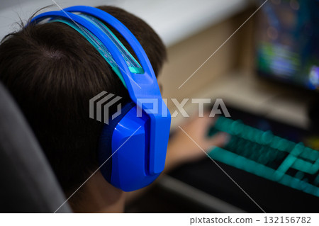 Close up of person wearing blue gaming headphones while playing game on computer. The vibrant colors of the headphones and keyboard highlight the immersive gaming experience 132156782
