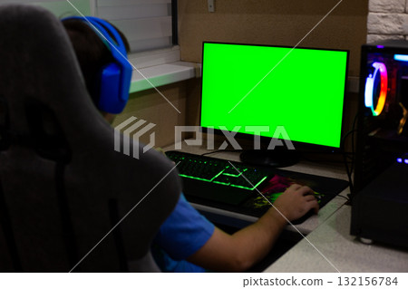 Person sits at gaming desk with backlit keyboard and mouse. The screen displays green screen, showing game setup with advanced computer hardware and gaming headset 132156784