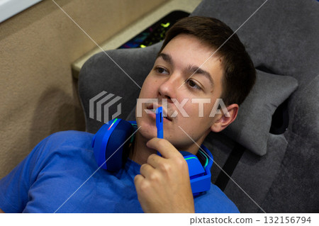Young gamer has removed his blue gaming headphones but is using microphone to communicate with other players, reflecting moment of interaction during intense gaming session Young gamer has removed his blue gaming headphones but is using microphone to communicate with other players, reflecting moment of interaction during intense gaming session 132156794