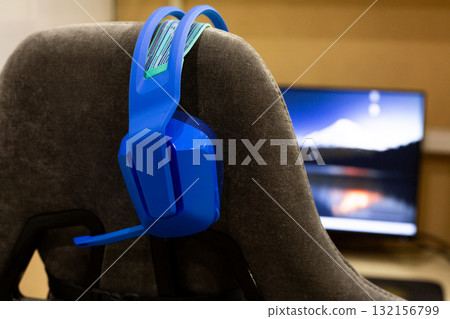 Blue gaming headphones hangs on back of gaming chair. The colorful design contrasts with the subtle background of computer screen, evoking sense of preparation for intense gaming action 132156799