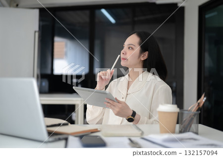 Businesswoman or office worker thinking while holding stylus and tablet sitting at table in office. Businesswoman or office worker thinking while holding stylus and tablet sitting at table in office. 132157094