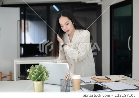 Businesswoman or female office worker is talking on the smartphone while leaning over working table. Businesswoman or female office worker is talking on the smartphone while leaning over working table. 132157105
