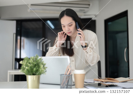 Businesswoman or female office worker is talking on phone while leaning to look at laptop on table 132157107