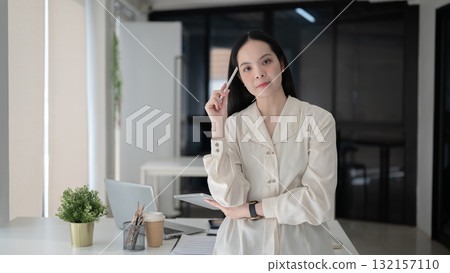 Female office worker holding tablet with stylus touching her head standing in front of working table Female office worker holding tablet with stylus touching her head standing in front of working table 132157110