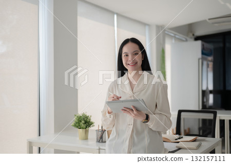 Female office worker is holding a stylus writing on tablet while standing in front of working table. Female office worker is holding a stylus writing on tablet while standing in front of working table. 132157111