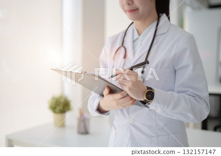 Female doctor with stethoscope writing with a pen on clipboard standing near table in diagnosis room Female doctor with stethoscope writing with a pen on clipboard standing near table in diagnosis room 132157147