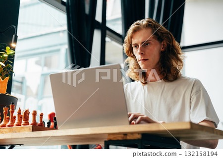 Young man focused on laptop while chess pieces sit nearby in a modern cafe Young man focused on laptop while chess pieces sit nearby in a modern cafe 132158194
