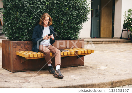 Young man sitting on bench using smartphone in urban area during daytime 132158197