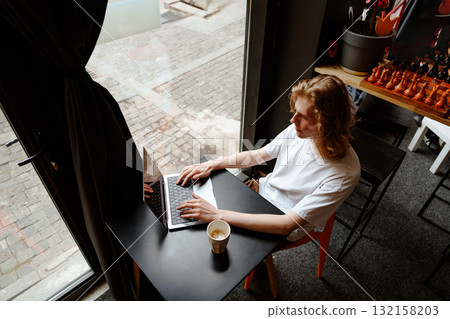 Young man works on laptop while enjoying coffee at a cozy cafe during daytime Young man works on laptop while enjoying coffee at a cozy cafe during daytime 132158203