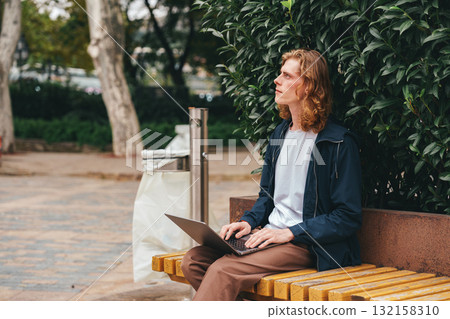 Young man works on laptop while sitting on a bench in urban park during a sunny day Young man works on laptop while sitting on a bench in urban park during a sunny day 132158310