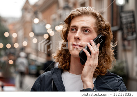 Young man talks on phone in busy city street during evening with lights in the background 132158323