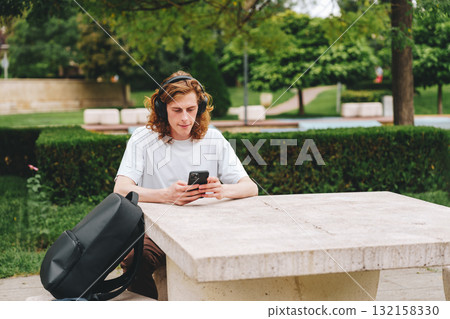 Young man using smartphone while sitting at a stone table in a park during daytime Young man using smartphone while sitting at a stone table in a park during daytime 132158330