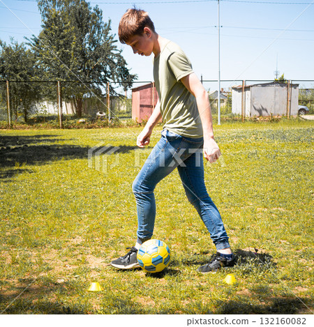 Teenager practicing soccer skills outdoors on a sunny day. He dribbles the ball near cones. 132160082