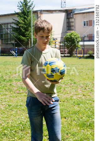 Teenager holding a soccer ball outdoors on a sunny day. He's near a building and some trees. 132160782