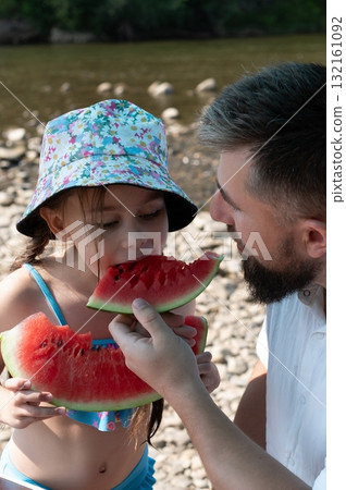 Father and daughter sharing a refreshing watermelon snack by the river on a summer day. Father and daughter sharing a refreshing watermelon snack by the river on a summer day. 132161092