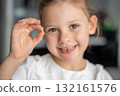 Smiling girl proudly holding her lost baby tooth between fingers. Emotional moment of childhood captured in a soft and cheerful portrait. 132161576