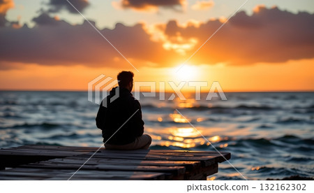 A tranquil scene at sunset. An individual sits alone on a dock, gazing out towards the vast ocean under a partly cloudy sky 132162302