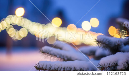 Winter evening scene with snow covered pine tree branch in foreground and blurred city lights in background, cozy christmas holiday and new year concept Winter evening scene with snow covered pine tree branch in foreground and blurred city lights in background, cozy christmas holiday and new year concept 132162723