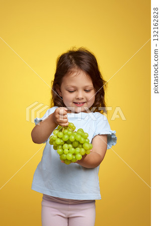 cute Caucasian little child girl holding bunch of green grapes on yellow background 132162828