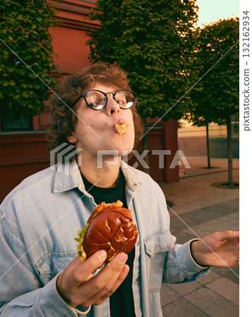 Cheerful young man eating burger on city street enjoying carefree moment 132162934