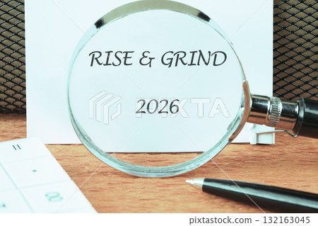 A close-up view of a motivational note displaying the words Rise Grind 2026. The magnifying glass emphasizes the message against a simple wooden background, inviting determination and focus 132163045