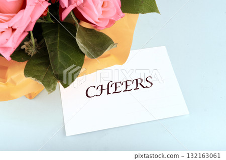 A vibrant bouquet of pink roses rests on a light blue surface. Next to it, a card with the word cheers adds a joyful touch to the scene, brightening the atmosphere 132163061