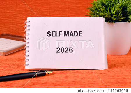 On a vibrant orange surface, a notebook titled Self Made rests beside a decorative plant and a sleek pen. This setup encourages creativity and inspires ambitious plans for the year 2026 On a vibrant orange surface, a notebook titled Self Made rests beside a decorative plant and a sleek pen. This setup encourages creativity and inspires ambitious plans for the year 2026 132163066