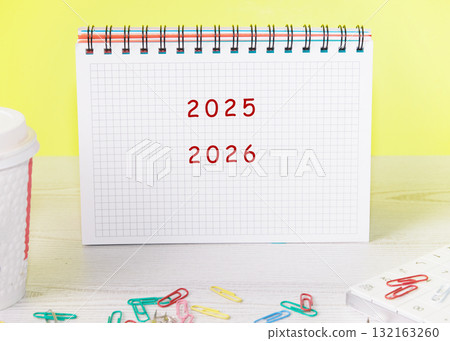 A calendar is opened to showcase the years 2025 and 2026. Colorful paper clips scattered around add a playful touch to this organized workspace against a cheerful backdrop 132163260