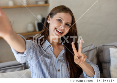 Smiling female taking selfie and showing v sign in living room Smiling female taking selfie and showing v sign in living room 132163417