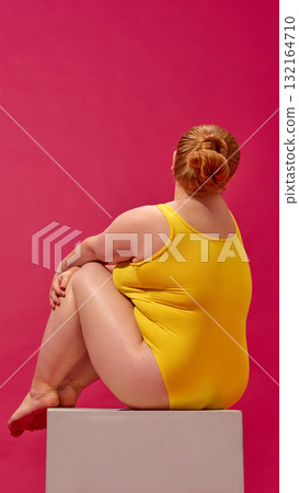 Rear view of plus-size woman sitting on cube in yellow swimsuit minimal composition 132164710