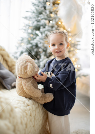 A young girl smiles while holding a teddy bear in front of a Christmas tree. A young girl smiles while holding a teddy bear in front of a Christmas tree. 132165020