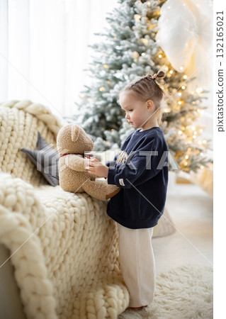 A young girl in a cozy setting interacts with a teddy bear near a decorated Christmas tree, creating a warm and festive atmosphere. A young girl in a cozy setting interacts with a teddy bear near a decorated Christmas tree, creating a warm and festive atmosphere. 132165021