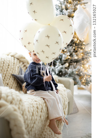 A young child smiles while holding balloons, with a Christmas tree in the background. 132165027
