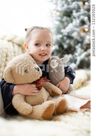 A young girl smiles while holding a teddy bear and a llama plush toy, with a Christmas tree in the background. 132165028