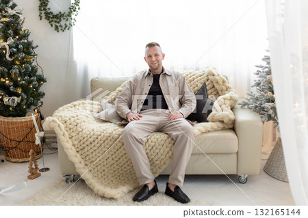 A man sits on a comfortable couch in a bright, cozy living room, surrounded by Christmas decorations. A man sits on a comfortable couch in a bright, cozy living room, surrounded by Christmas decorations. 132165144
