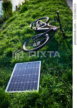 Pink electric mountain bike lies on lush green grass, connected to solar panel for charging. Bicycle beside paved path, showcasing bike's rugged tires and sleek design in outdoor environment. 132166223